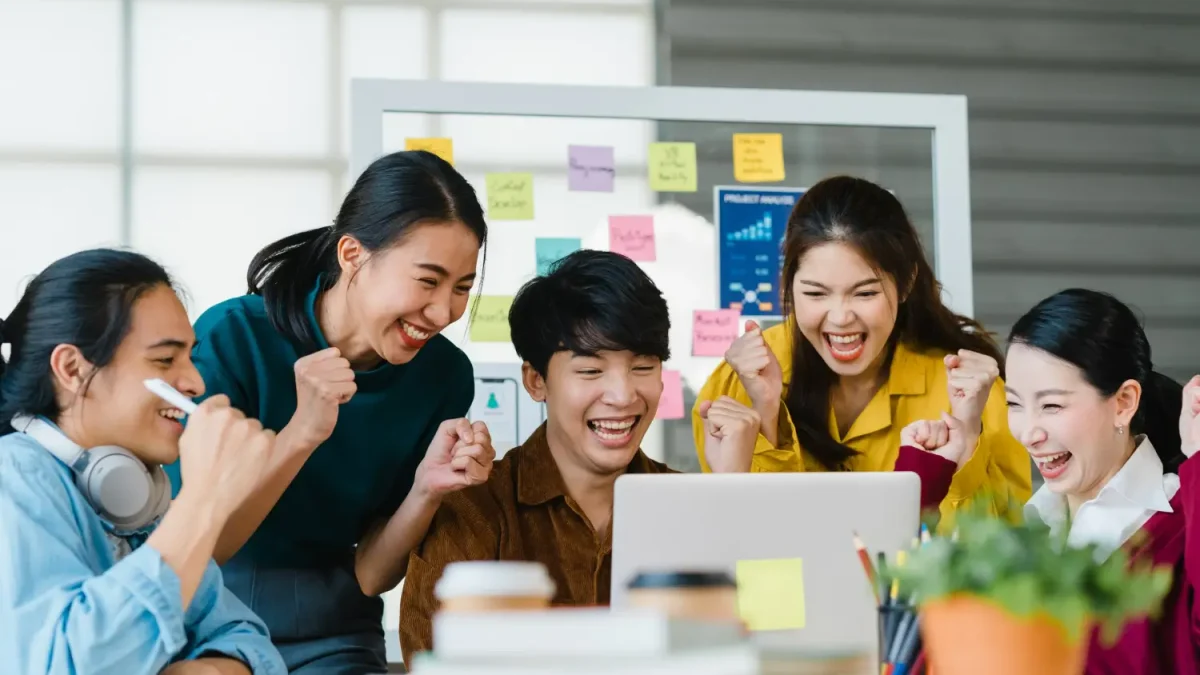 Tim Digital Agency yang kompak merayakan keberhasilan kampanye di depan laptop, menunjukkan efisiensi dan sinergi tim agensi dibanding in-house.