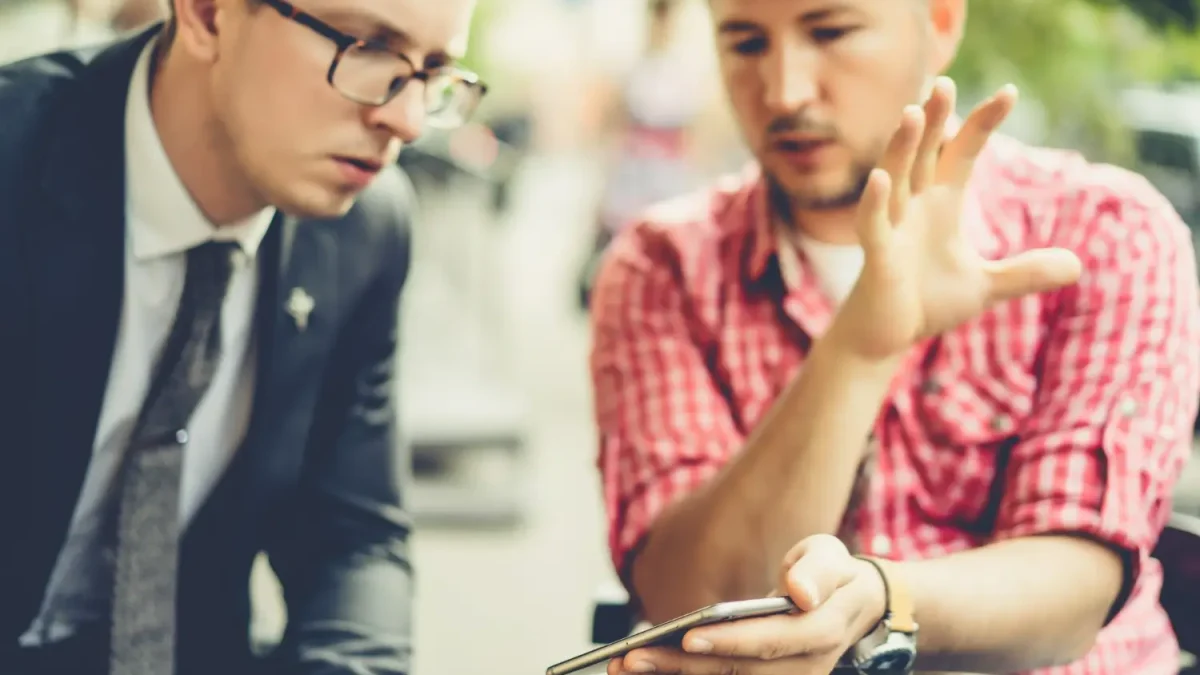 Dua orang pria sedang berdiskusi serius sambil menunjuk layar smartphone, mengilustrasikan proses analisis sentimen dan percakapan audiens dalam strategi social listening.