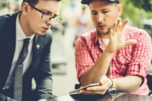 Dua orang pria sedang berdiskusi serius sambil menunjuk layar smartphone, mengilustrasikan proses analisis sentimen dan percakapan audiens dalam strategi social listening.