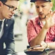 Dua orang pria sedang berdiskusi serius sambil menunjuk layar smartphone, mengilustrasikan proses analisis sentimen dan percakapan audiens dalam strategi social listening.