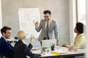 Manajer proyek sedang mempresentasikan strategi di papan tulis kepada timnya, merencanakan penawaran sponsorship event yang komprehensif dan berbasis data untuk menarik sponsor potensial.
